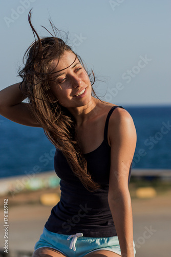 Photo of beautiful young brunette woman relaxing on the resort under the hot sun. Summer and vacation concept. Portrait of Caucasian suntanned girl with sea and blue sky in background.
