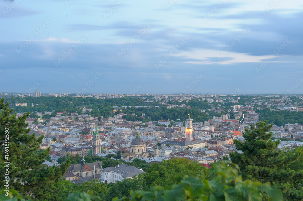 Obraz premium View of Lviv city center from a hilltop at dusk, with blurred sky and city lights glowing, trees in the foreground.