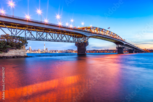 Auckland Harbour Bridge at Twilight, with its lights on and a red reflection in the harbour.
