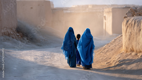 Two women in traditional blue veils and a child walking along a dusty road near mud-brick houses, afternoon sunlight, rural life, cultural heritage Middle East, traditional clothin