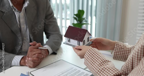 A real estate agent presents a property sale contract to a client, discussing agreement details with a model house on the document, symbolizing home buying, selling, and real estate investment.