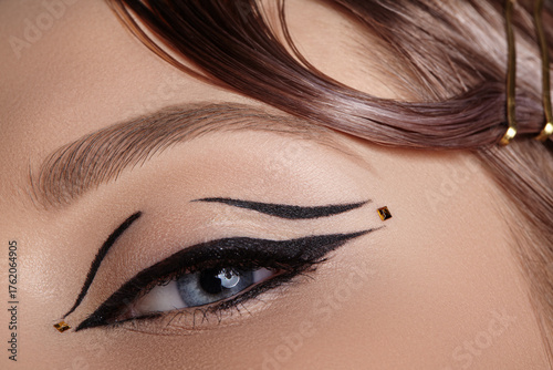 Close-up macro of a closed female eye with black winged eyeliner and tiny gold dot embellishment, soft lighting highlighting lash curve and eyelid decoration for editorial beauty use