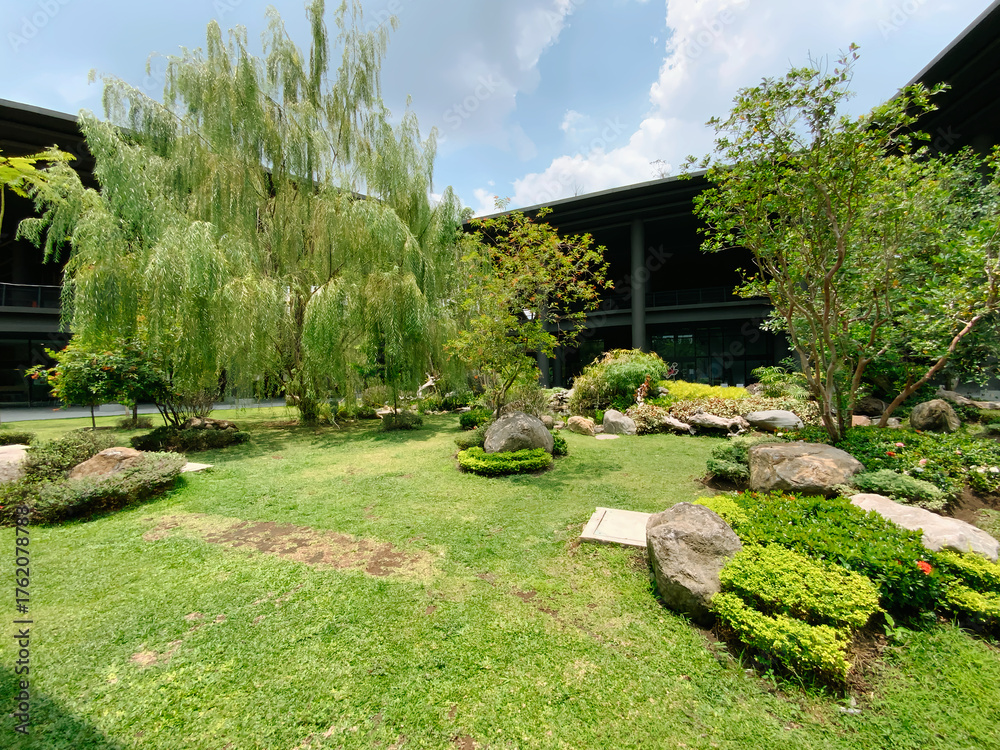 Fototapeta premium Serene modern courtyard garden with lush green grass, weeping willow, and natural rock landscaping offers tranquil business retreat vibes, located in Yogyakarta Indonesia.