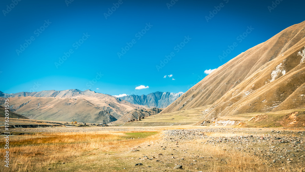 Fototapeta premium Panoramic view of Truso Valley in Georgia on a sunny autumn day. Golden grasslands and majestic Caucasus Mountains under a deep blue sky create a peaceful natural scene