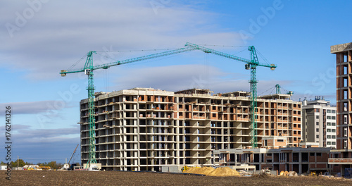 A wide shot of a multi-story residential building under construction, featuring two prominent tower cranes against a bright blue sky, symbolizing urban development, real estate growth, and modern civi