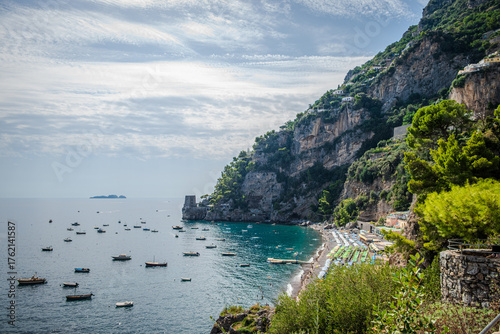 A coastal view of the Amalfi Coast from Positano, Italy, featuring boats on the Tyrrhenian Sea, steep cliffside mountains, and a faint island visible in the distance