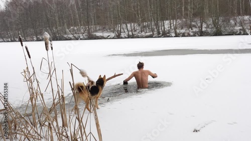 Brave man takes an ice plunge in a frozen lake while his loyal dog plays nearby. Concept of winter swimming, healthy lifestyle, and extreme sports. High quality 4k footage