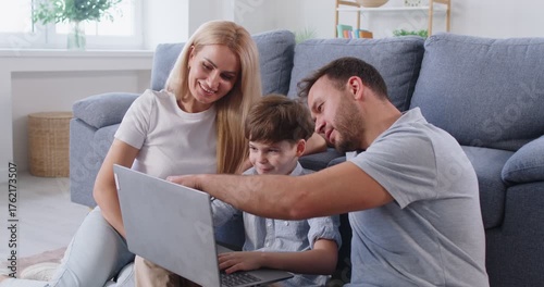 Little boy sitting with laptop on floor with parents beside him showing something on screen. Happy father, mother and son spending leisure time together, exploring or learning something new as family.