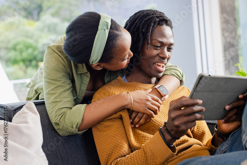 African American couple sitting on dark grey sofa using tablet in living room with green foliage