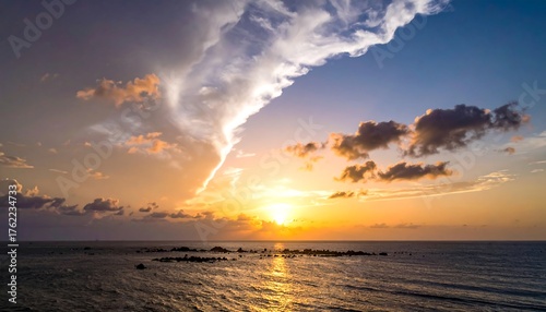 An aerial photograph captures a golden sunset over a calm ocean. Fluffy clouds illuminate as the sun dips below the horizon. Rocks jut from the sea