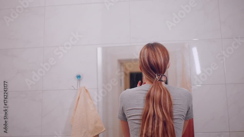 Back view of young girl standing in front of mirror observing face with calm expression, hair clipped neatly behind, soft indoor lighting, simple tiled bathroom