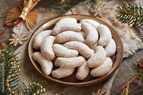Homemade Christmas cookies called Kipferl or vanilla crescents coated in sugar on a brown plate with decorated spruce tree
