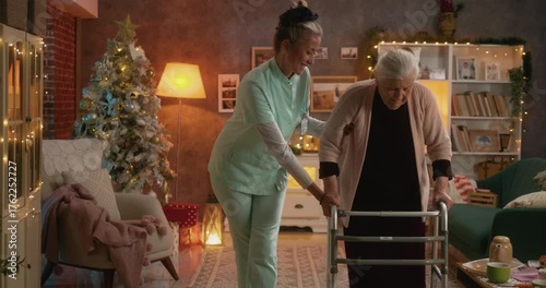 Kind caregiver supporting elderly woman walking with walker in festive living room during Christmas, showing care and gentle assistance.