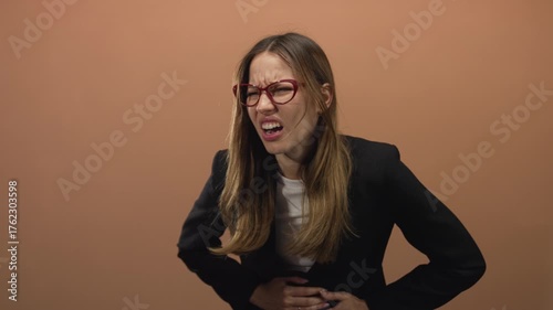 Young hispanic woman grimacing and clutching stomach with hands in peach studio; stomach pain discomfort.