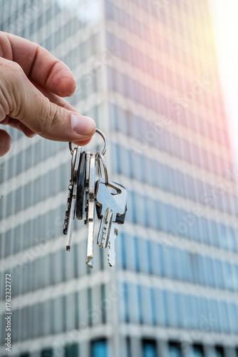 Real Estate Keys Held by Hand in Front of Modern Office Building. Close-up shot of a hand holding several metal keys in front of a sleek modern glass skyscraper