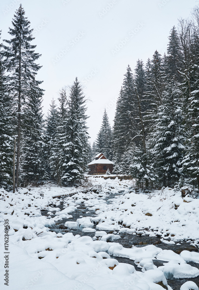 Naklejka premium Rustic wooden cabin by a mountain stream in a snowy winter forest.