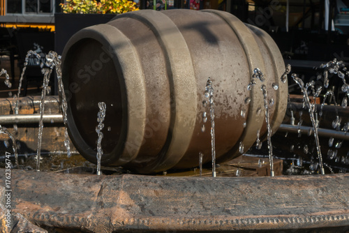 Water fountain with wooden barrel and circular jets in outdoor setting. Rustic barrel fountain with metal stand and water jets in public plaza with tables and chairs under natural daylight.