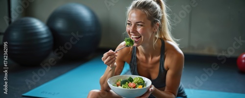 Fit woman eats healthy salad after gym workout. Female athlete rests on yoga mat enjoying fresh vegetables. Nutrition for strong body and mind.
