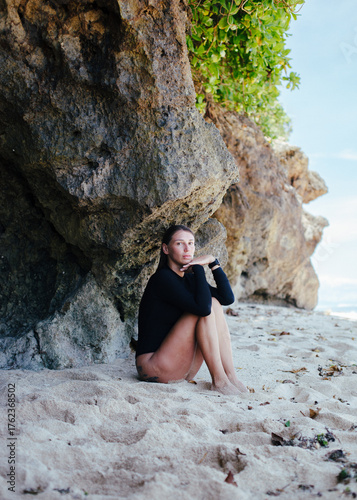 Portrait of young woman in black surf suit sitting on sandy beach in Bali, Indonesia