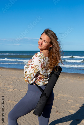Woman smiling on beach carrying yoga mat over shoulder