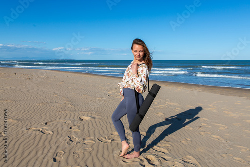 Woman standing barefoot on beach carrying yoga mat in sunlight