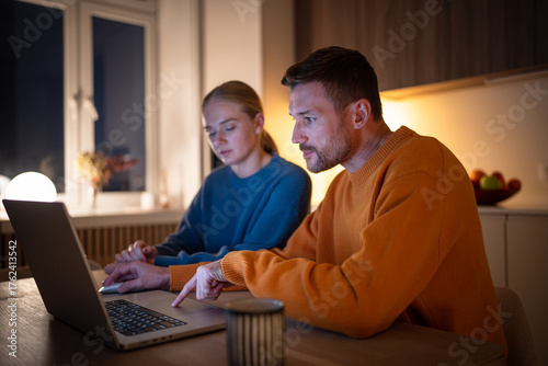 Photography Exhausted young couple telecommuting, browsing and working on laptop in kitchen at night