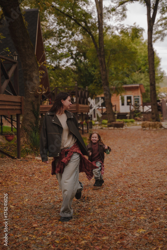 Mother and daughter run around the autumn yard near cozy A-shaped houses. The girls appear to be dressed in leather jackets, blue jeans and plaid shirts. Autumn atmosphere. Vertical photo.