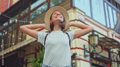 Happy young traveler girl in straw hat with backpack raising hands in relaxing gesture enjoying city street view near hotel on vacation
