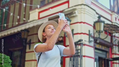 Happy young traveler girl in straw hat with backpack filming landmarks and street architecture on phone in tourist city