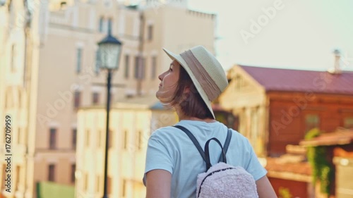 Thoughtful pretty tourist girl in straw hat with backpack in casual clothes looking around tourist city street enjoying travel