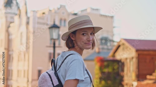 Young tourist girl in straw hat with backpack back view turning with smile looking at camera on sunny street during travel through ancient city