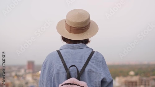 Traveler girl in straw hat with backpack looking at city from summer terrace height back view city view from bird's eye view