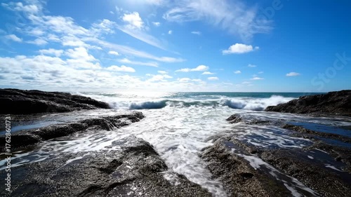 Ocean's Embrace: Witness the raw power and beauty of the ocean as waves crash against rugged rocks, all under a bright blue sky, the sun illuminating the scene