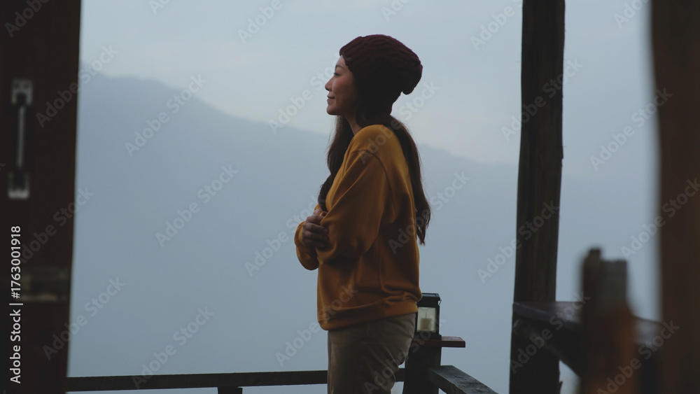 Fototapeta premium A woman standing on wooden balcony with mountain views on foggy morning outside the window