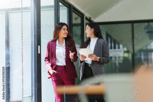 Two Asian businesswomen discussing work while walking outside a modern office building, one holding a digital tablet