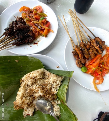 A delicious Indonesian meal featuring satay skewers, spicy tomato sambal, and nasi bakar wrapped in banana leaves, served on white plates for a traditional and flavorful dining experience.