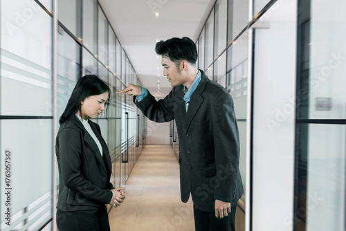 Fotografija Angry Asian male manager scolding or blaming his sad, submissive female employee in a modern office hallway, depicting workplace conflict and harassment