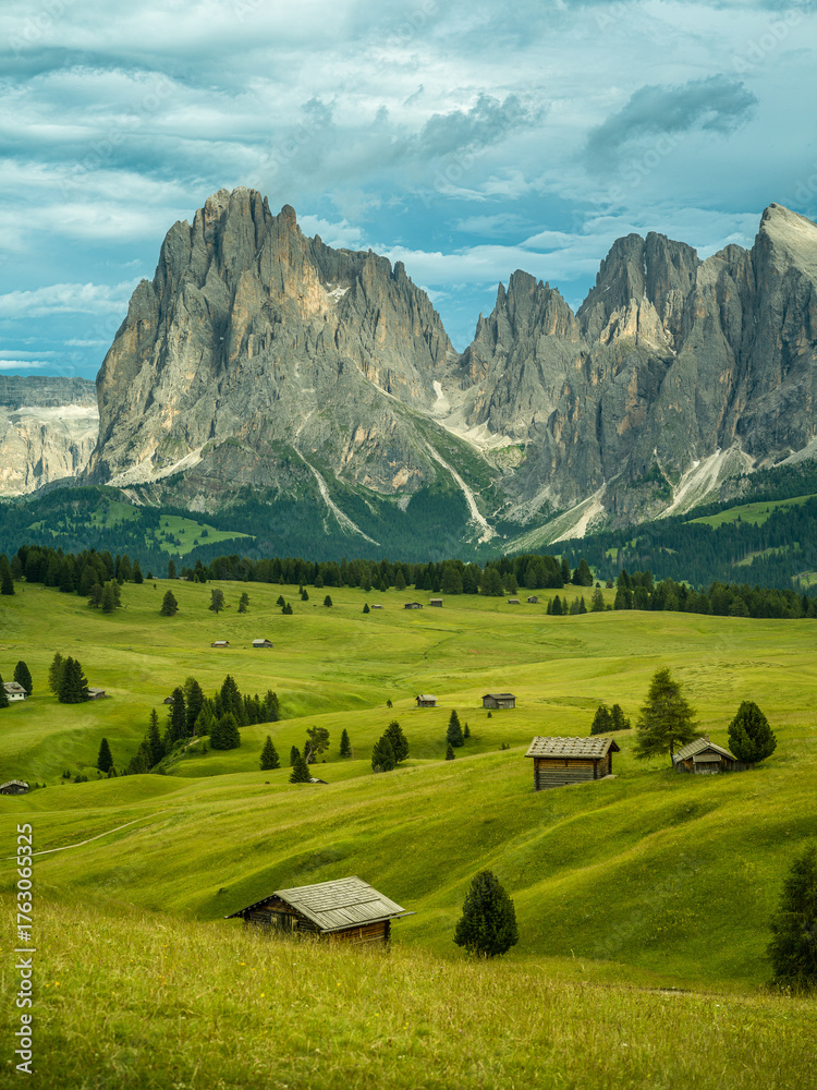 Fototapeta premium Dramatic landscape in the Dolomites, Italy. Green alpine meadows, wooden huts, and majestic mountains under a mix of sunlight and storm clouds — pure natural harmony and power.