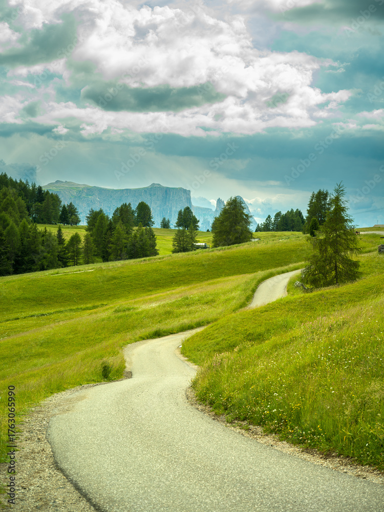 Obraz premium Hiking path in Dramatic landscape in the Dolomites, Italy. Green alpine meadows, wooden huts, and majestic mountains under a mix of sunlight and storm clouds — pure natural harmony and power.