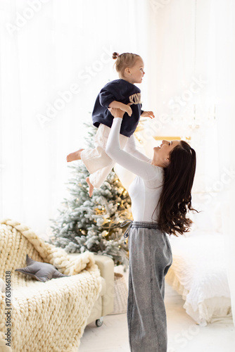 A mother joyfully lifts her child in the air, creating a heartwarming moment indoors. A Christmas tree is in the background.