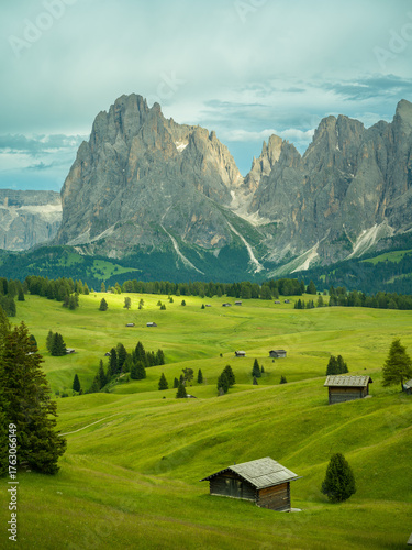 Dramatic landscape in the Dolomites, Italy. Green alpine meadows, wooden huts, and majestic mountains under a mix of sunlight and storm clouds — pure natural harmony and power.