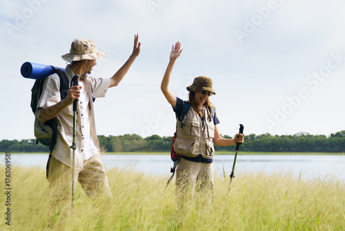 Asian man and woman happily high-fiving each other after reaching their hiking destination in a beautiful grassy meadow by the lake, celebrating success, friendship, and outdoor adventure in nature.