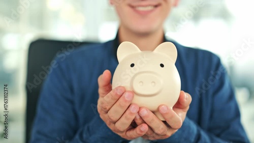 Cheerful man holds a beige piggy bank in an office, wearing a blue shirt. Smiling male embodies the concept of financial planning, savings, and future investments with happiness.
