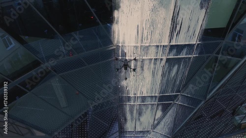 aerial view of Industrial drone spraying foam and water while cleaning glass facade of modern office building with sunset reflections