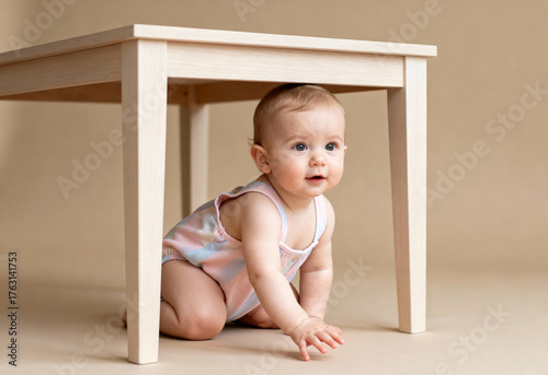 Curious baby in pastel outfit crawling under wooden table on neutral background with natural light and playful expression