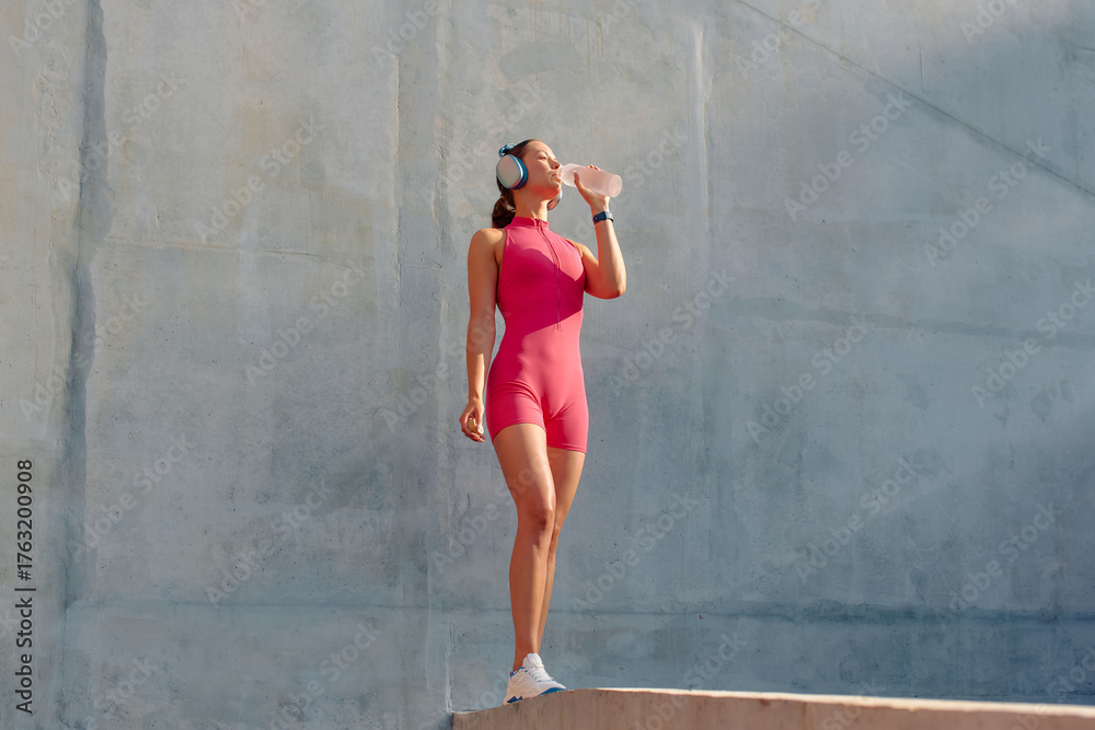 Fototapeta premium Outdoor sports activity featuring a woman wearing a pink outfit and headphones drinking water