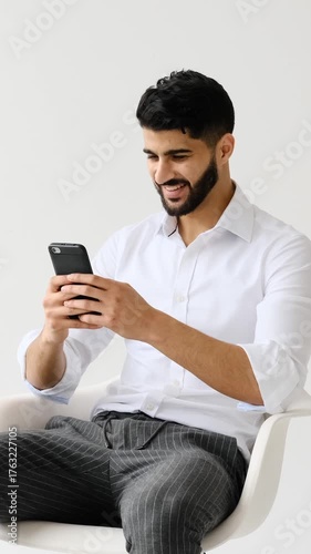 A young adult businessman sits in a chair against a white background, smiling as he types on his smartphone and takes a selfie.