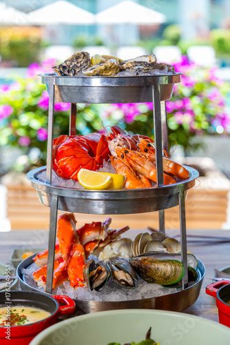 Seafood platter served on ice, featuring oysters, lobster, shrimp, crab, and clams. Outdoor setting with vibrant flowers in background, offering fresh luxury dining experience.