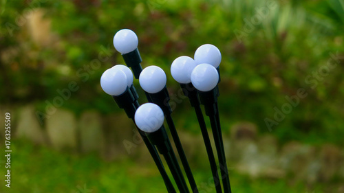 A close-up of modern garden lights with white spherical bulbs standing against a green blurred background. The image shows minimalistic outdoor design and peaceful natural atmosphere.