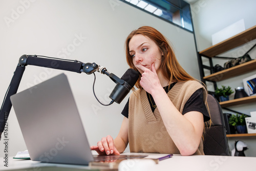 Cuadro en lienzo A young woman sits at a desk with a laptop, speaks into a cardioid mic on a boom arm, and touches her chin in a modern home studio with shelves, plants, and soft light